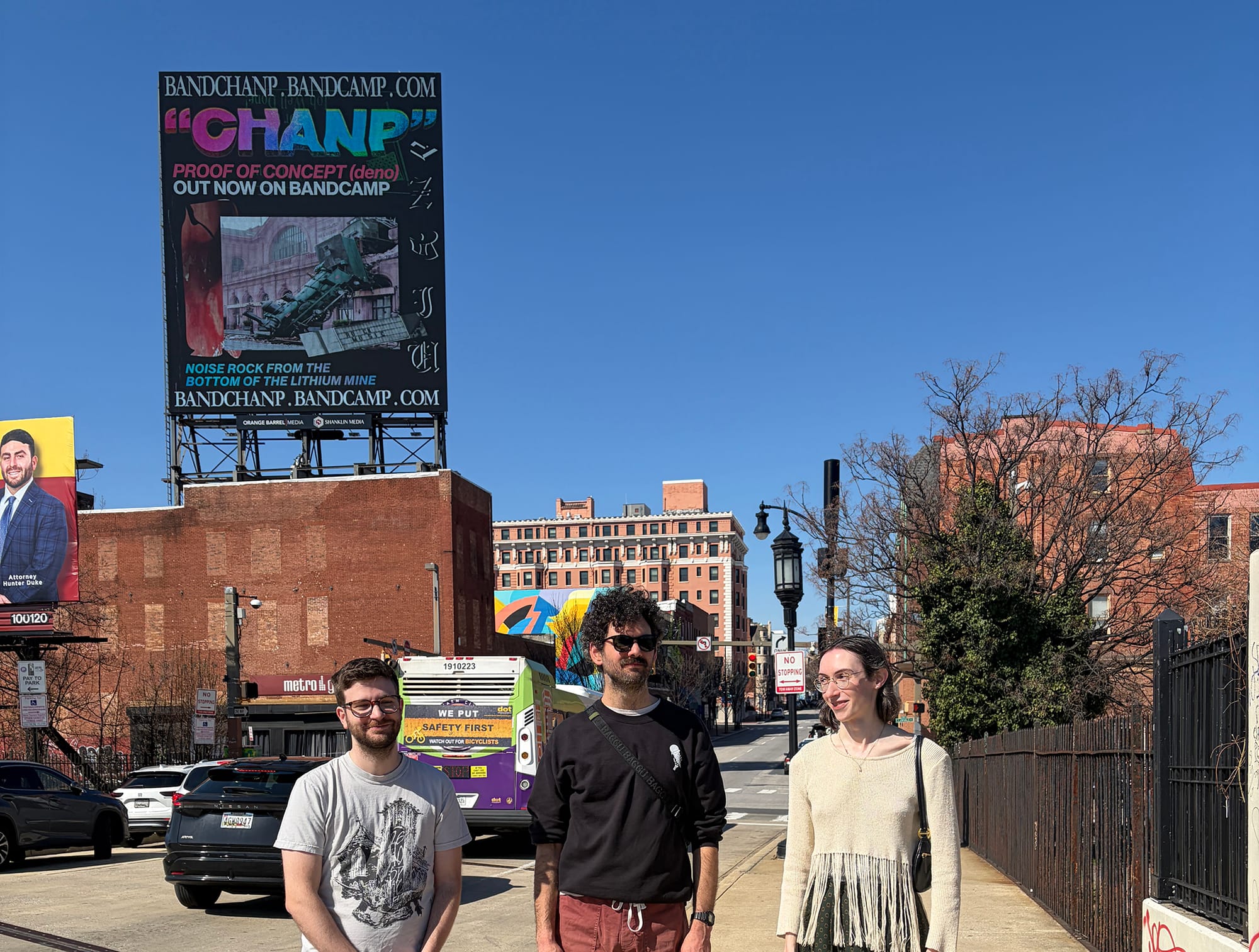 The band Chanp stands in front of a big billboard that says "Chanp Proof of Concept (deno) out now on Bandcamp. Noise rock from the bottom of the lithium mine"