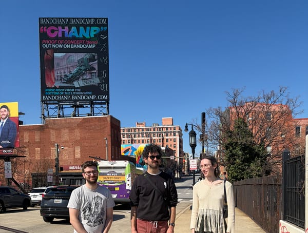 The band Chanp stands in front of a big billboard that says "Chanp Proof of Concept (deno) out now on Bandcamp. Noise rock from the bottom of the lithium mine"